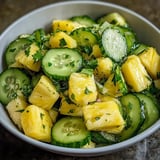 A dish with chunky pineapple and sliced cucumber inside.